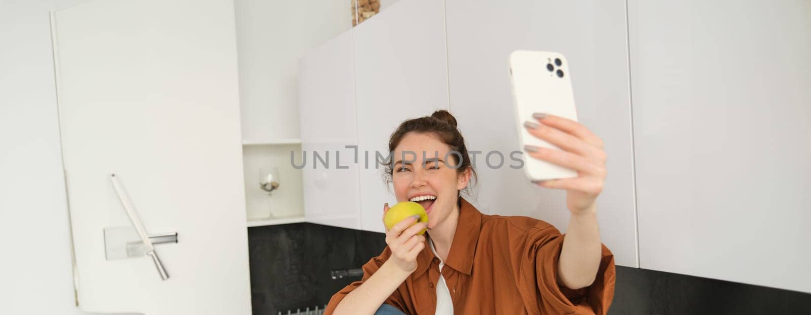 Beautiful young woman, lifestyle blogger records video of herself, takes selfie while biting an apple, sitting on kitchen counter, using smartphone by Benzoix