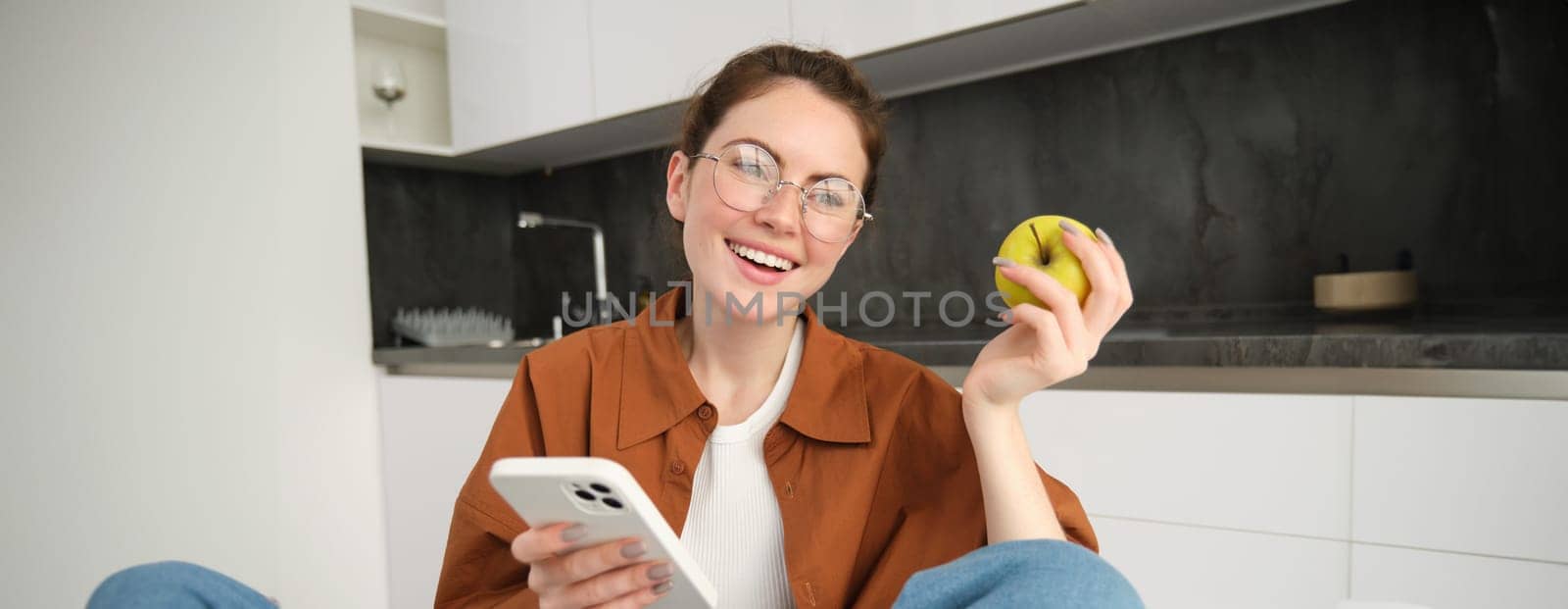 Portrait of happy, beautiful young woman in glasses, sitting with mobile phone, eating apple, laughing and smiling, relaxing, browsing social media application by Benzoix