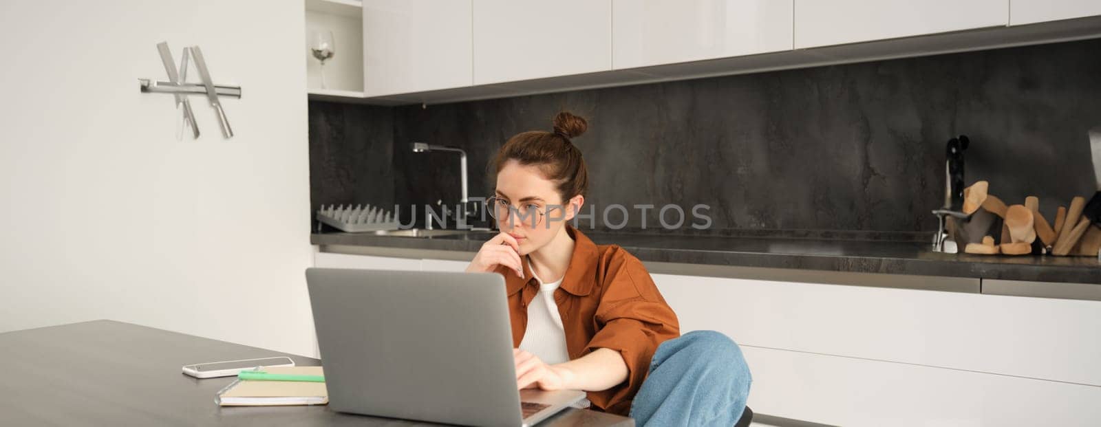 Portrait of woman sitting at home with laptop. Businesswoman managing her own business remotely from her kitchen. Student looking at computer screen, studying, doing her homework by Benzoix