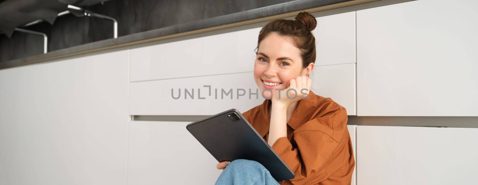 Portrait of young beautiful woman sitting on kitchen floor with digital tablet, browsing news feed, social media app on gadget, smiling and looking happy.