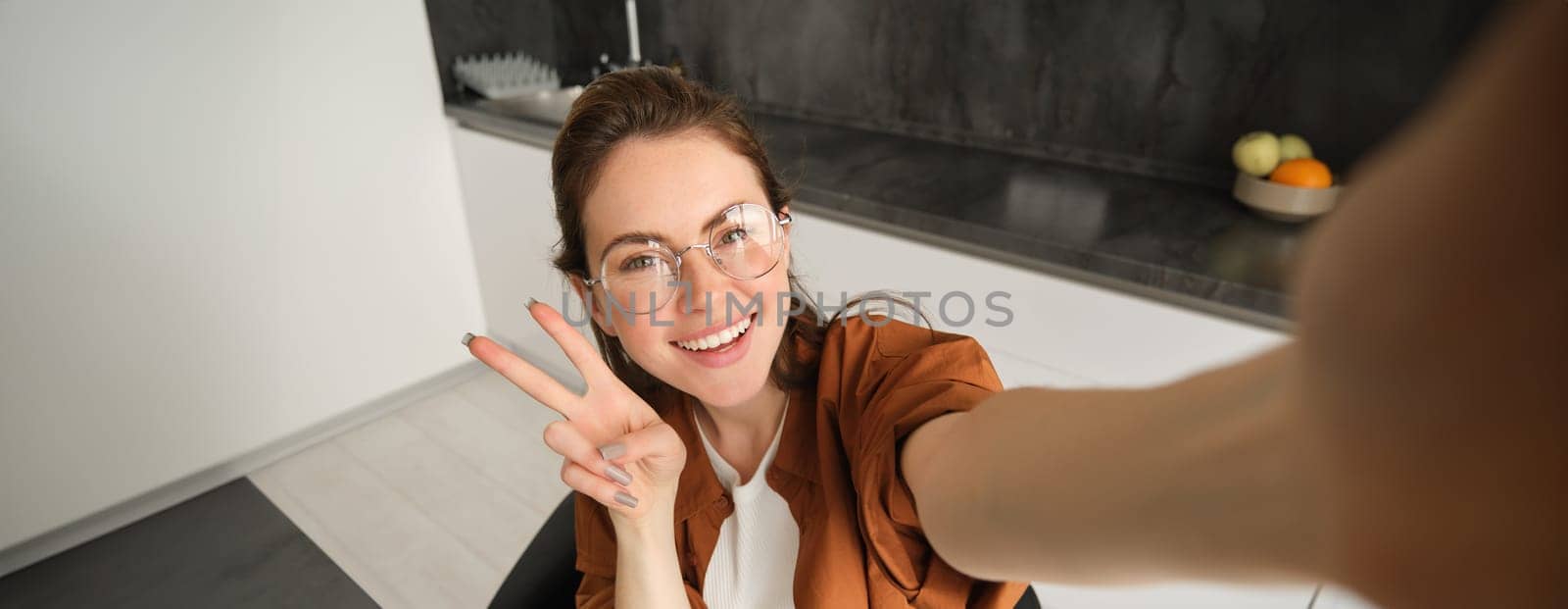 Selfie of stylish young woman, lifestyle blogger capturing her weekend at home, posing for photo, extends hand and holds smartphone, smiles and shows v-sign by Benzoix