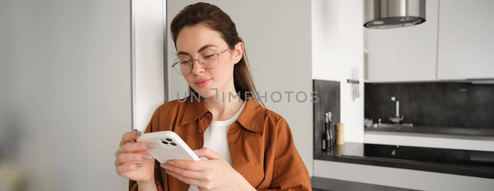 Portrait of young woman at home, checking news or messages on smartphone, online shopping, confirm delivery on mobile phone app, leaning on wall, standing in living room by Benzoix