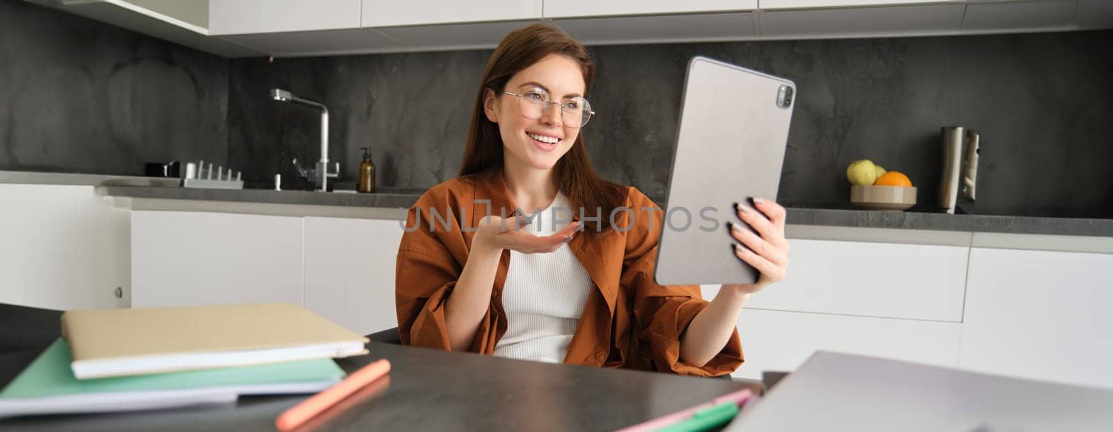 Portrait of young woman working from home, sitting in kitchen and talking with team, has online meeting, using digital tablet to join webinar, having conversation by Benzoix