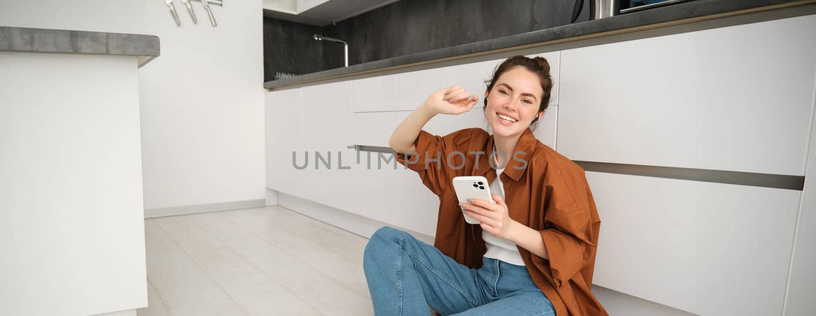 Smiling modern woman at home, sits on kitchen floor with mobile phone, wearing wireless black headphones, listening music or podcast, watching videos on smartphone app by Benzoix