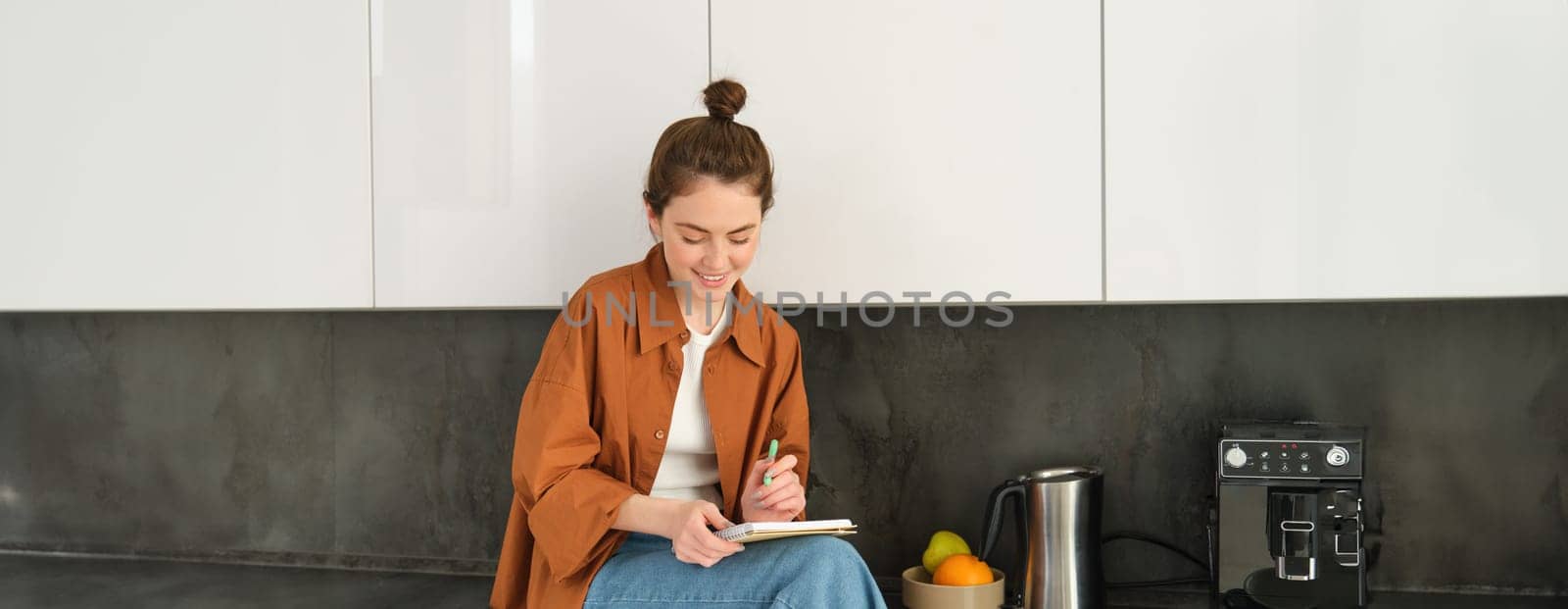 Portrait of stylish young woman, sits at home in the kitchen and makes notes, writes down recipe, checks her list with house errands, doodling or drawing in notebook.