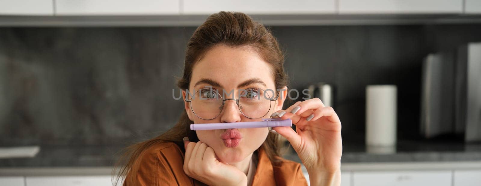 Close up portrait of funny, silly young woman, playing with pen, holding pencil on top of her lip and grimacing, sitting in kitchen.
