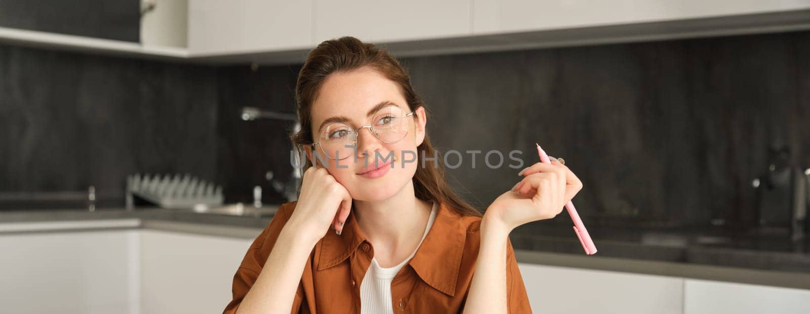 Close up of dreamy smiling woman, sitting in kitchen with pen, looking outside window, writing, making notes by Benzoix