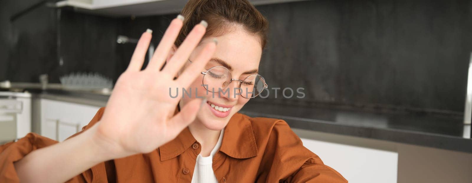 Close up portrait of cute young woman, covers, puts hand as block from camera and smiling, sitting in kitchen, laughing and smiling by Benzoix