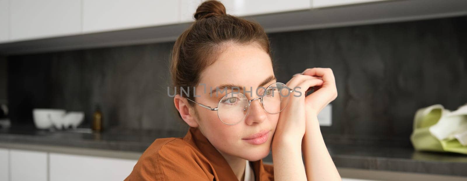 Close up portrait of gorgeous young woman in glasses, looking thoughtful and relaxed, posing sitting in kitchen, smiling with tenderness by Benzoix