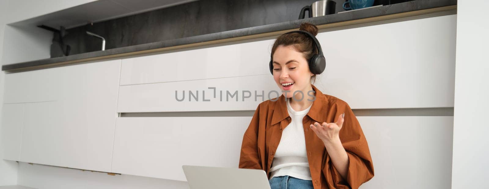 Young woman, freelancer joins online chat, student studying at home, sits on floor with laptop and headphones.