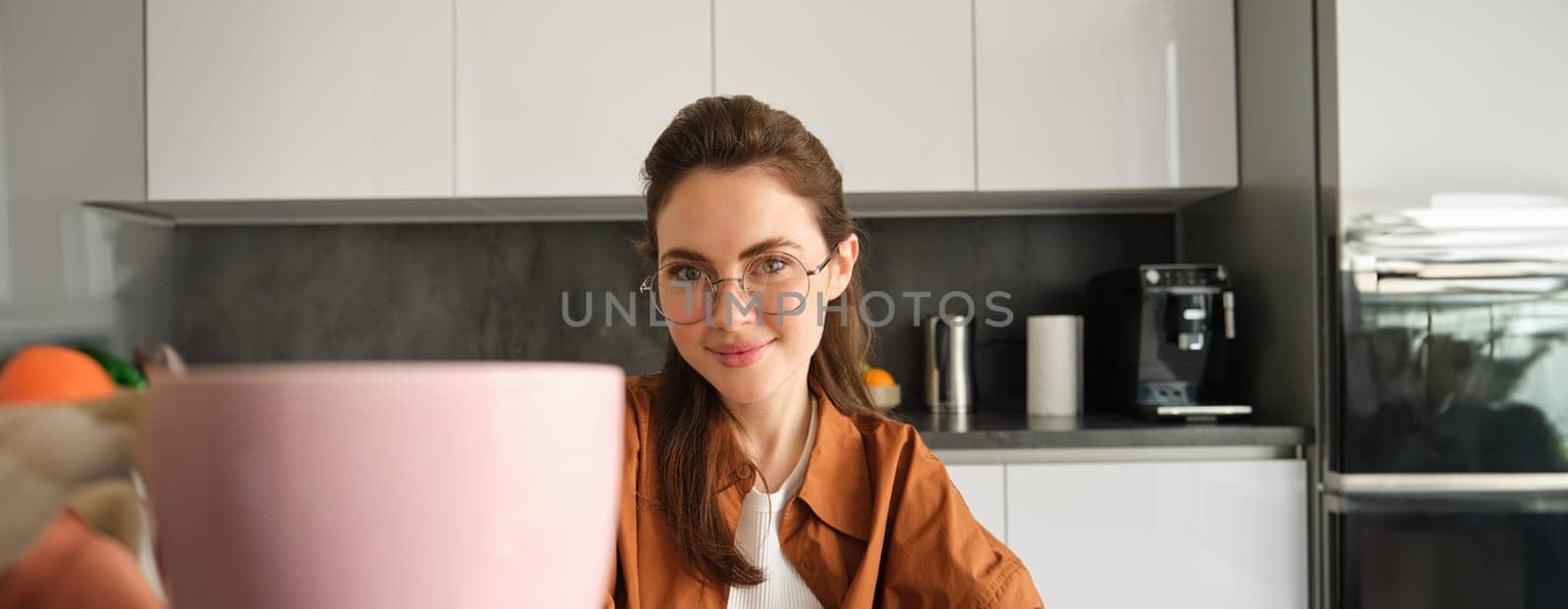 Close up portrait of beautiful young woman, giving you cup of coffee, inviting for tea, sitting in kitchen and smiling.