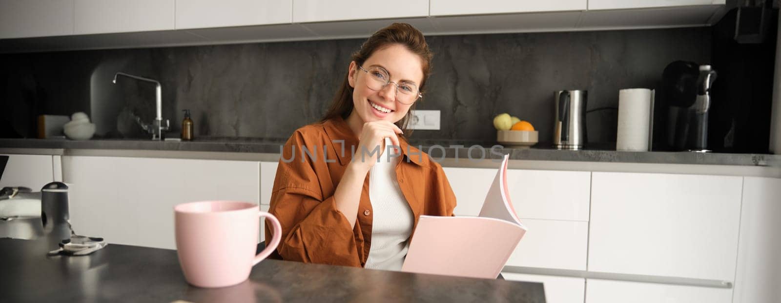 Portrait of beautiful young woman working from home, freelance tutor preparing for lesson. Student sitting in kitchen with folder, reading documents.