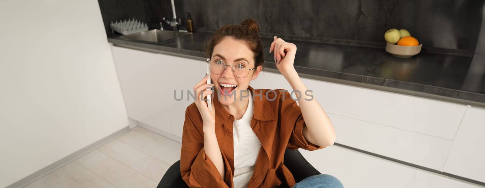 Portrait of woman receiving great news over the phone call, celebrates and jumps on chair from excitement during telephone conversation.