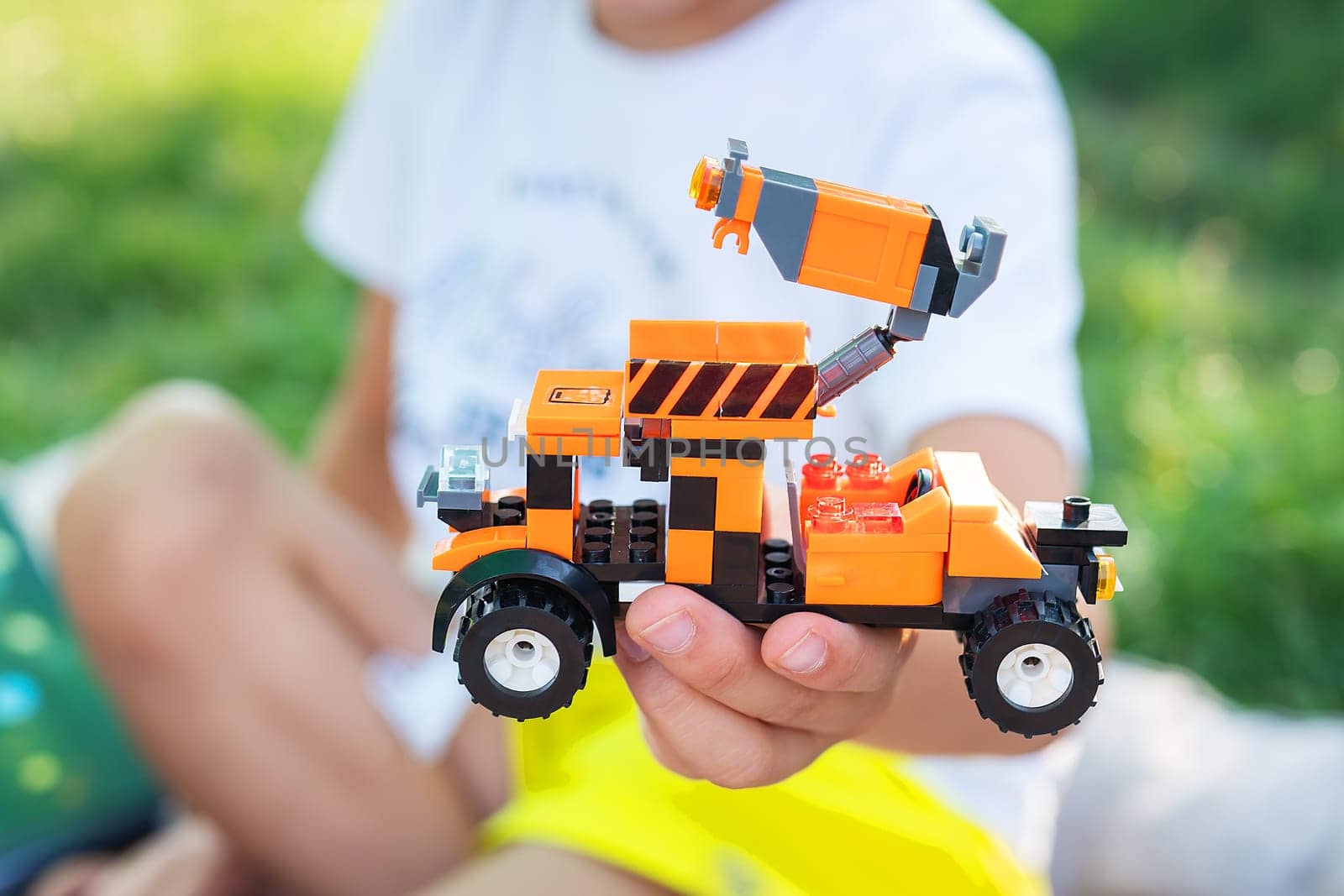 A child s hands holding a toy truck made of building blocks. The truck is orange and black with stripes on the side. The child is sitting on a grassy lawn in a park on a sunny day