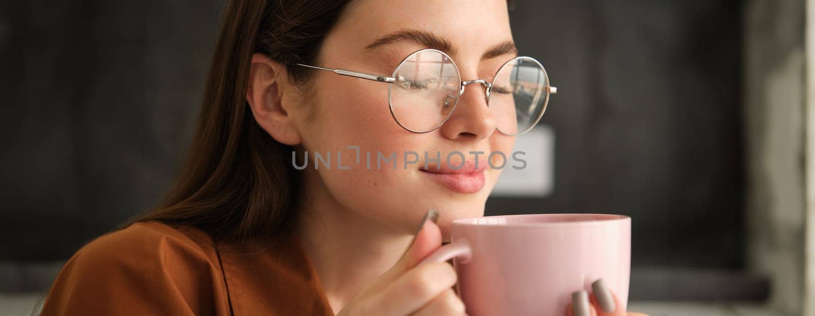Close up portrait of beautiful smiling woman, drinks her morning cup of coffee, enjoys tasty aroma, smelling her drink with pleased face by Benzoix