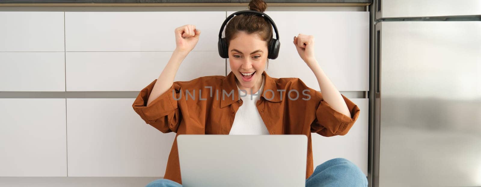 Excited young woman, programmer achieves goal, triumphing, winning on laptop, sitting in headphones, dancing with hands, celebrating victory.