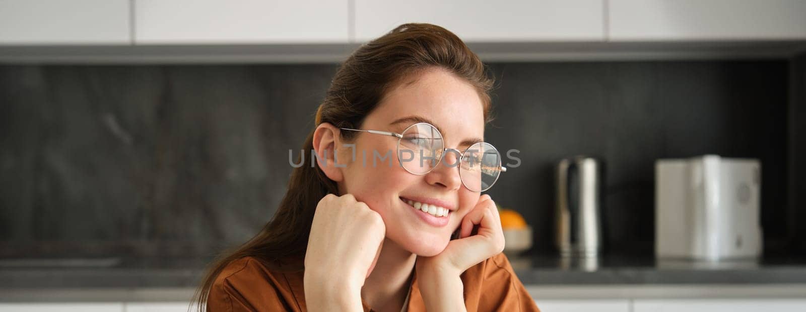 Portrait of lovely, happy young woman, wearing glasses, sitting in kitchen with dreamy, thoughtful face expression, spending time at home.
