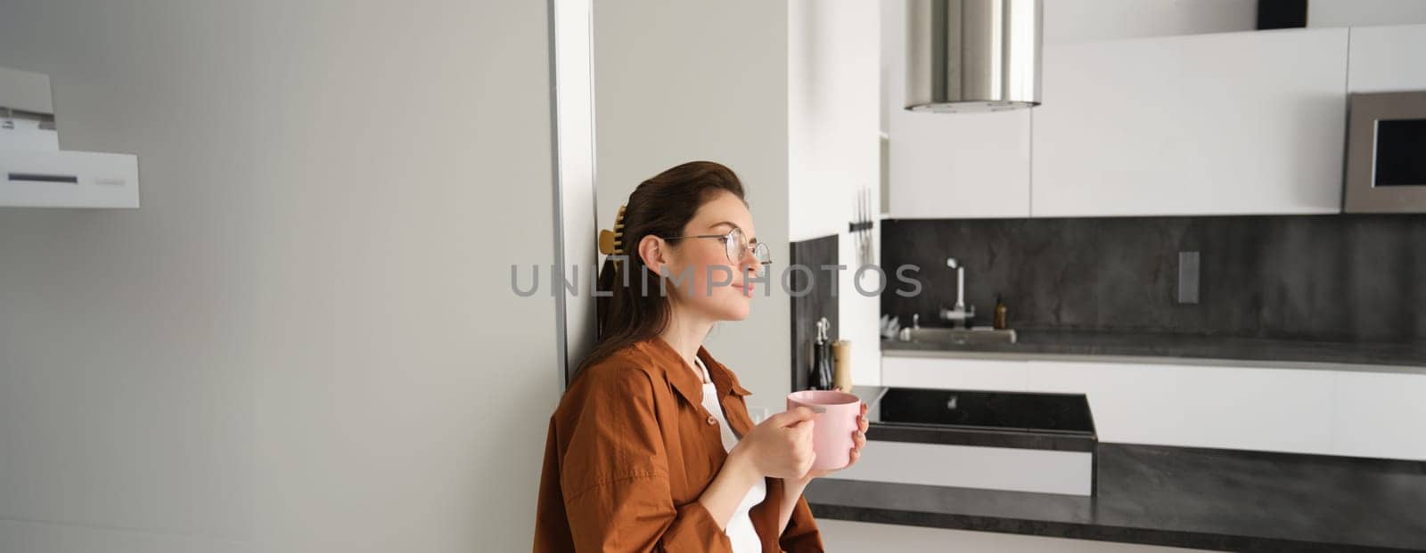 Portrait of happy young woman at home, holding cup of tea, leaning on wall, smiling, drinking coffee from pink mug by Benzoix