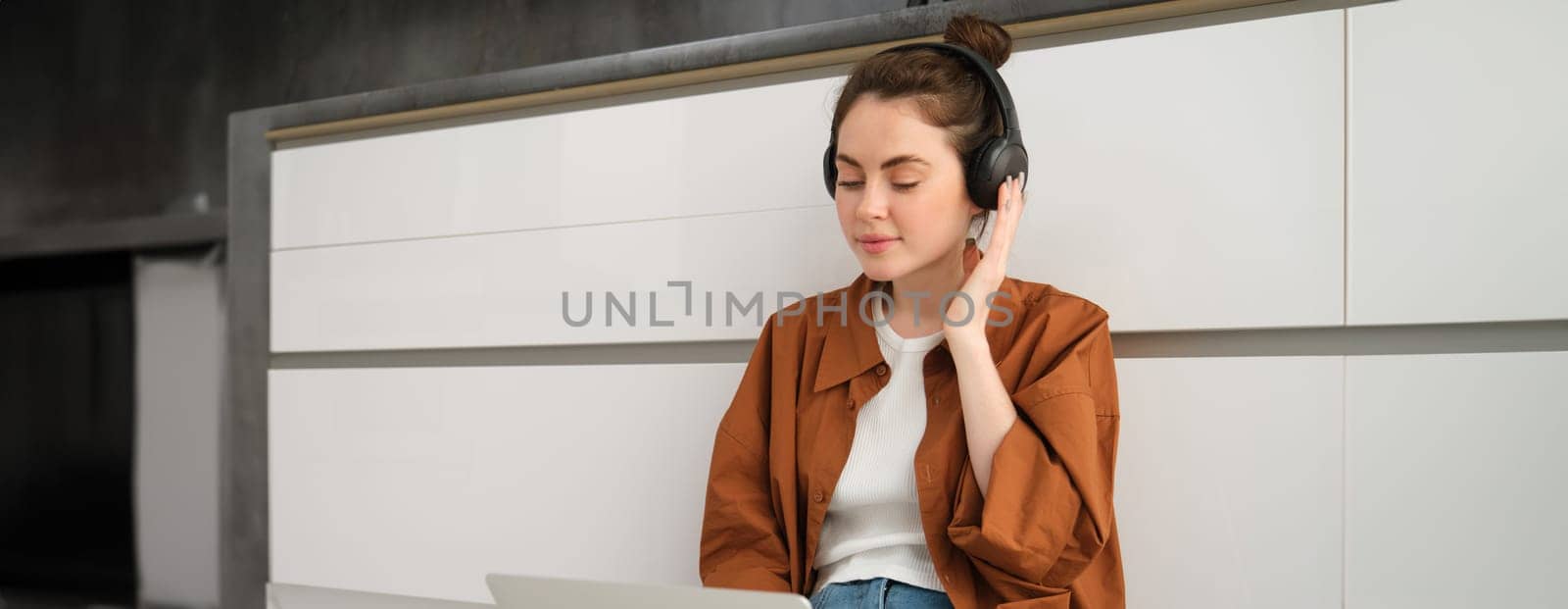 Young woman, student doing homework on laptop, sitting on floor, listening to music in headphones and working from home.
