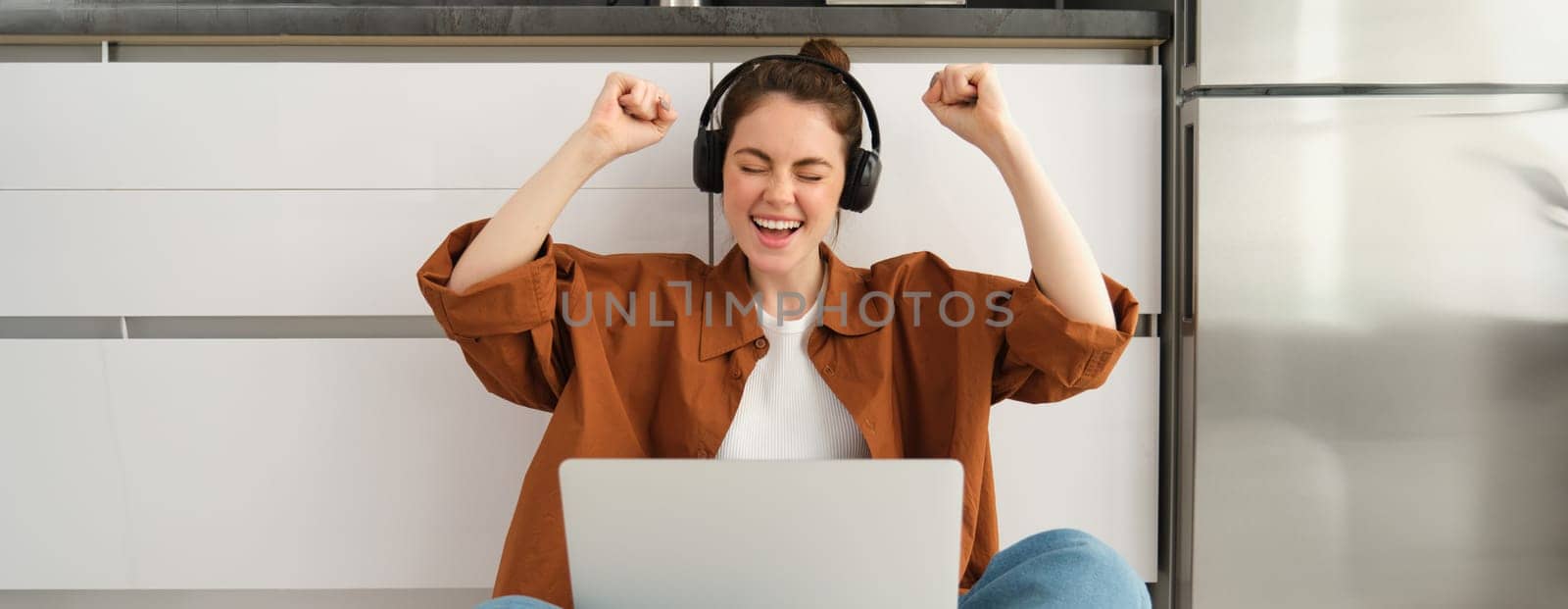 Enthusiastic young woman achieves goal, sits on floor with laptop and headphones, screams with excitement, wins in game on computer, celebrates victory, triumphs.