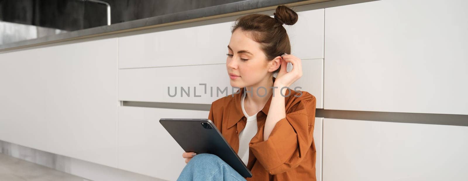 Portrait of cute young woman, freelancer working from home, sitting on kitchen floor with digital tablet and smiling.