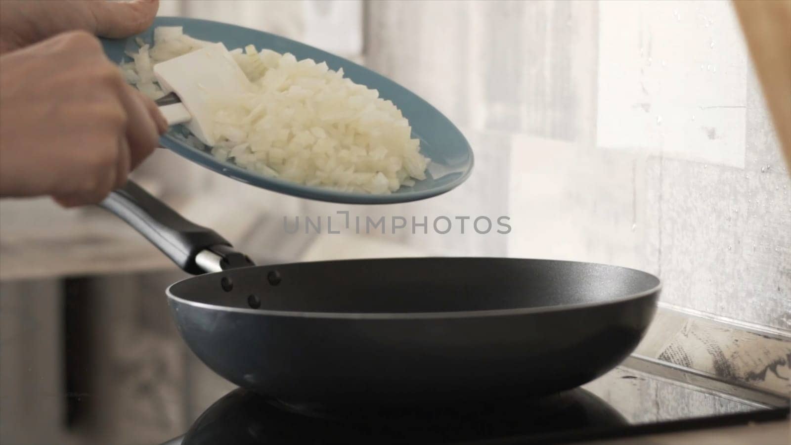Side view of woman hands putting cut onion into pan to fry. Close up of woman putting fresh onion from the glass plate into the pan in the kitchen.