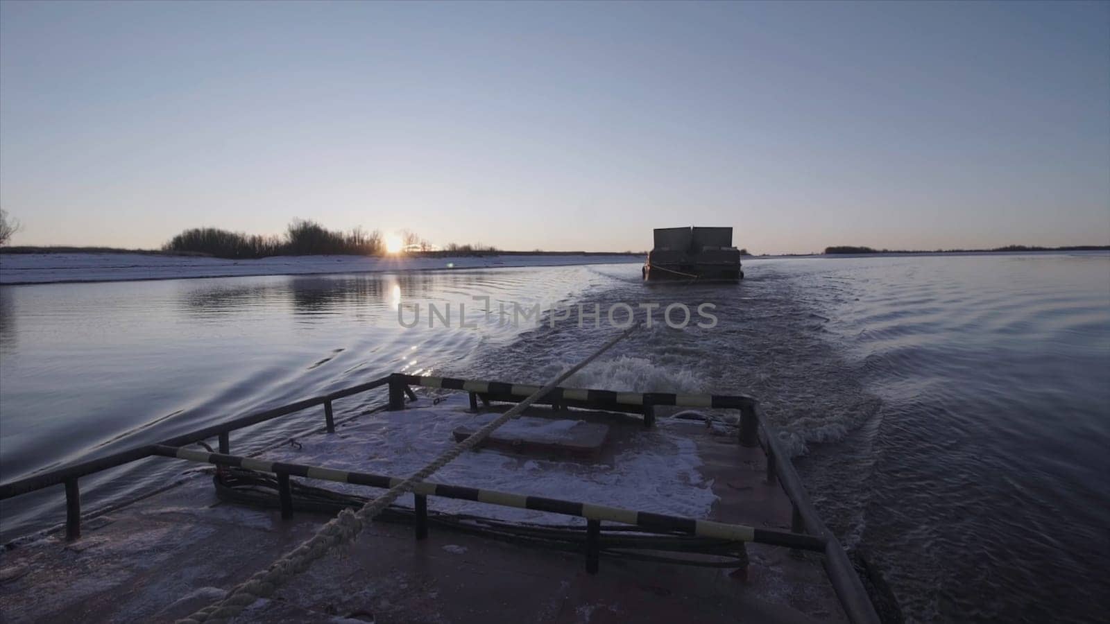 View of floating barge with cable on river. Clip. Floating barge behind ship near shore with snow on sunny winter day. Barge on cable floats behind boat on river.