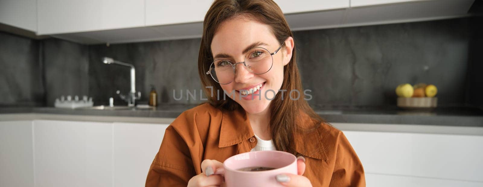 Close up portrait of beautiful woman in glasses, drinks tea, warms her hands with hot cup, smiling and looking happy at camera, sitting in kitchen.