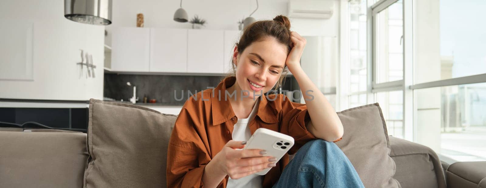 Portrait of happy young brunette woman sitting on sofa with smartphone, reading notification, messaging on mobile phone, resting on couch at home, smiling while looking at device screen.