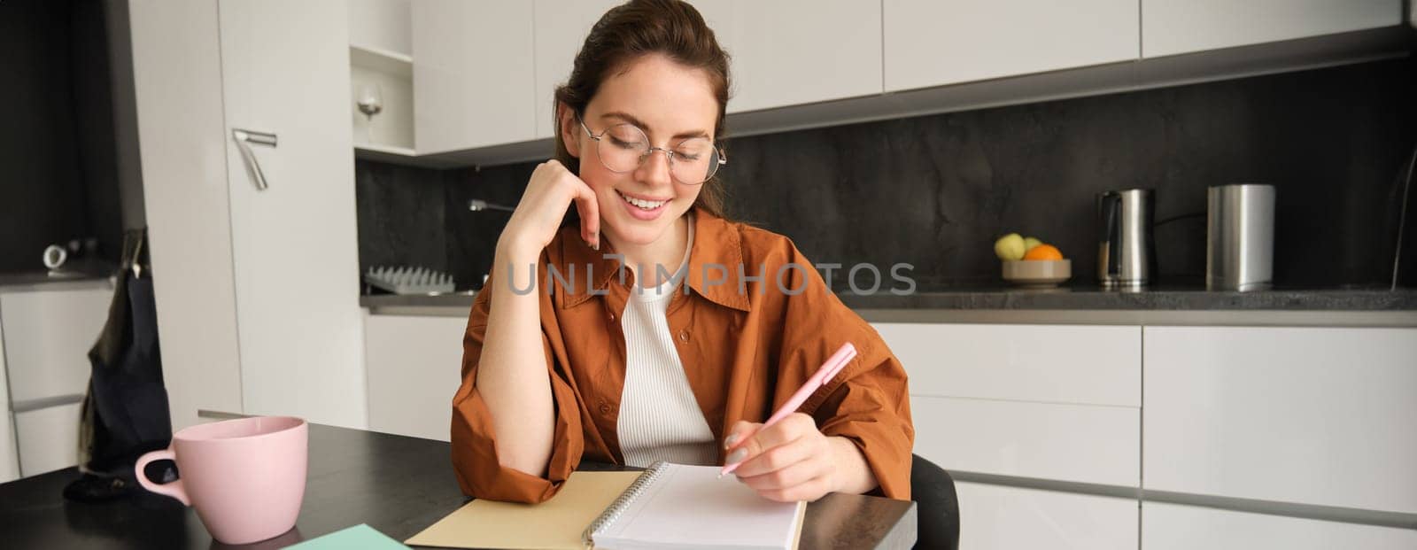 Portrait of modern young woman in glasses, student sitting at home and doing homework, writing down information in notebook and smiling, working on project.