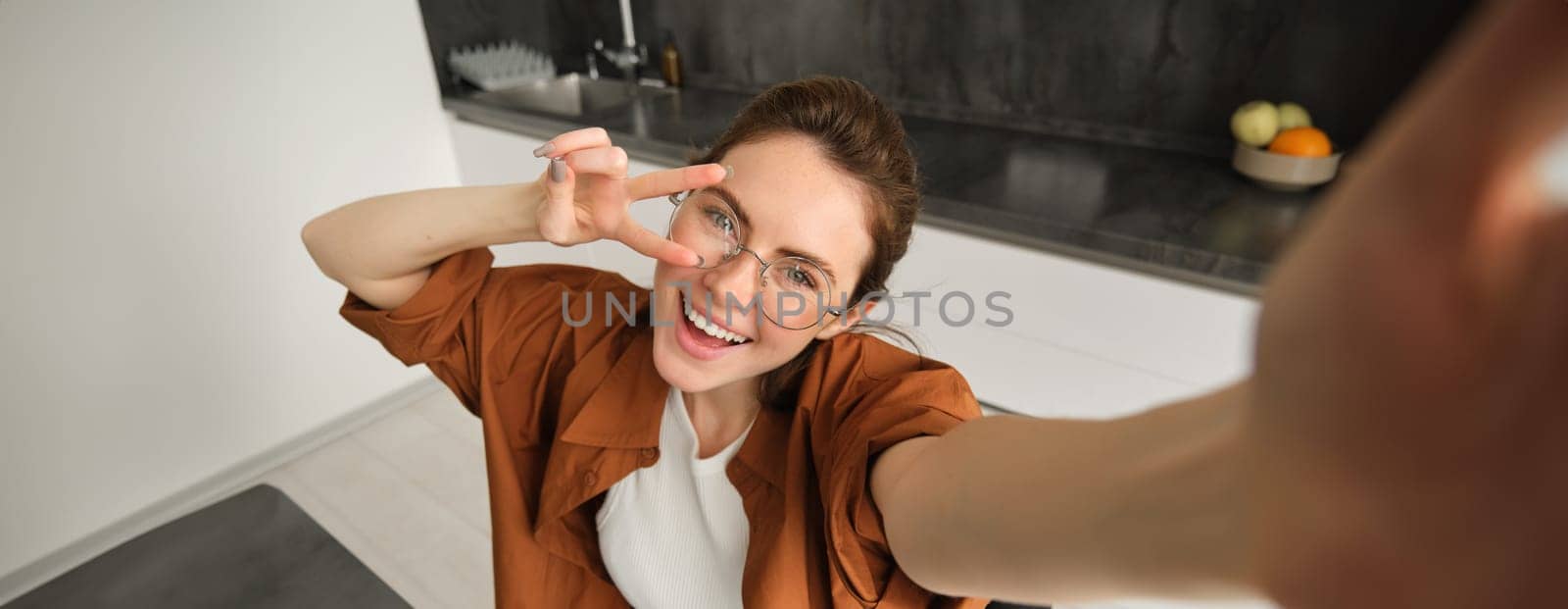 Selfie of happy young and carefree woman, taking photo on mobile phone with extended hand, posing and smiling, sitting in kitchen in glasses.