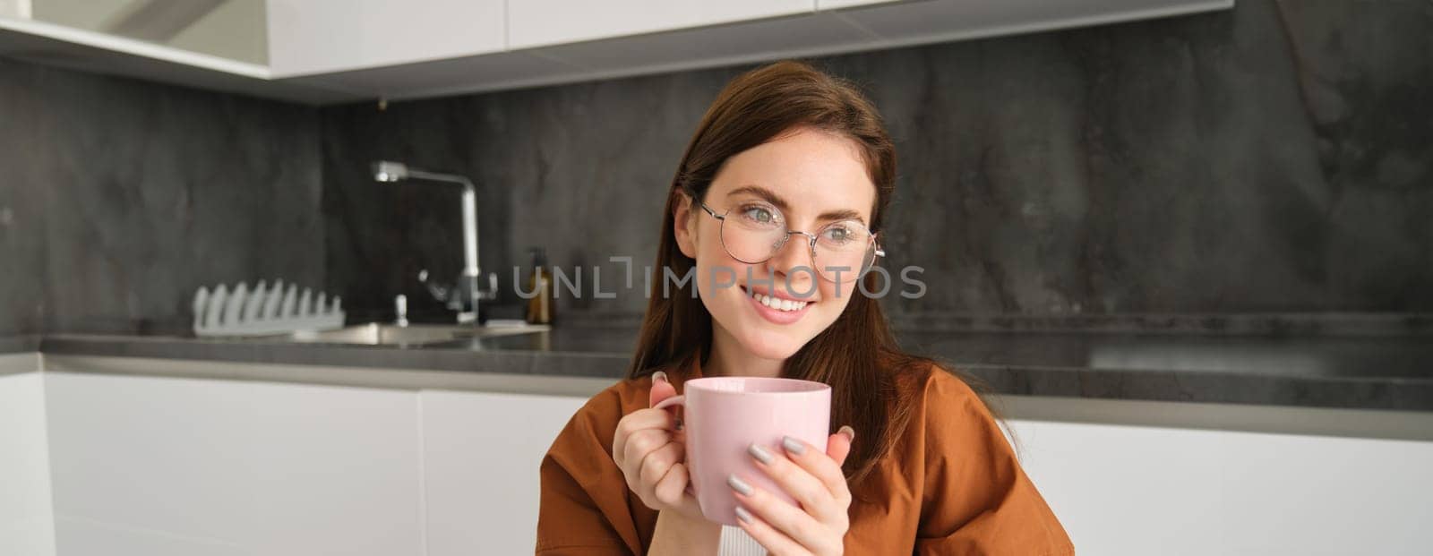 Woman drinking hot coffee at home. Thoughtful young woman drinking a cup of tea while thinking. Pretty girl with sweater relaxing at home while drinking purifying herbal tea by Benzoix