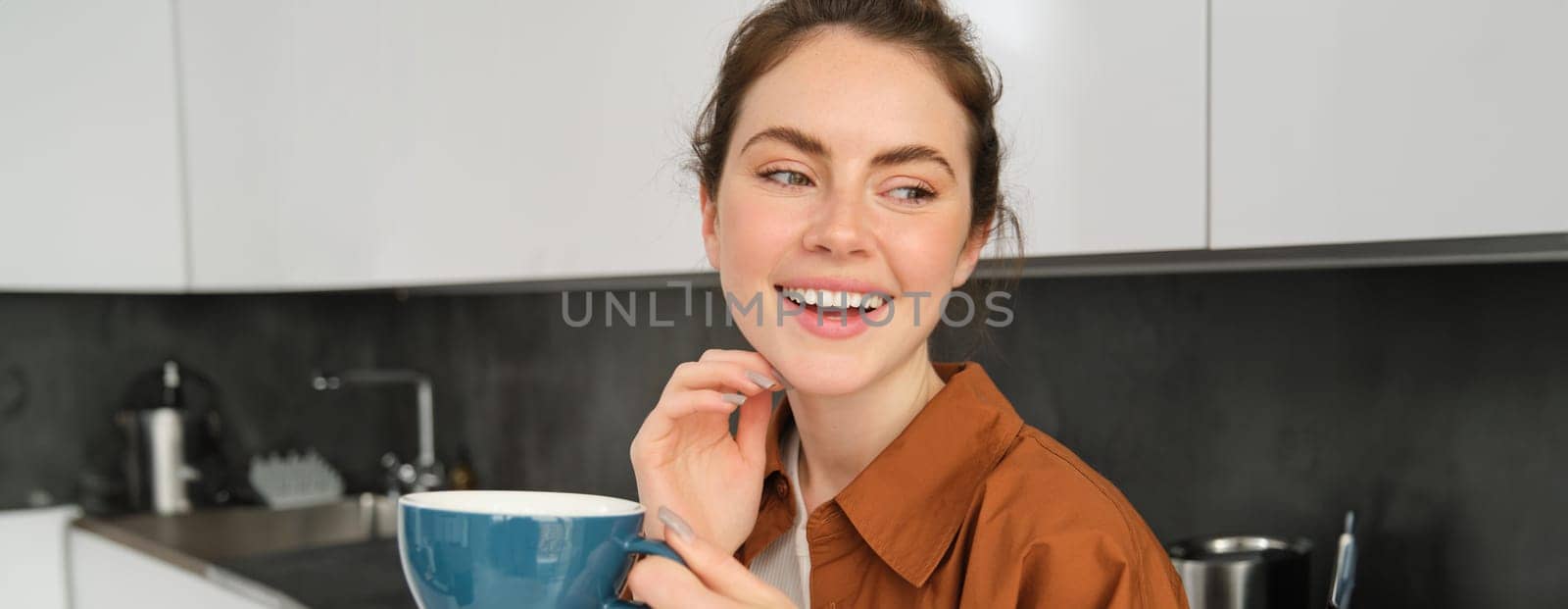 Close up portrait of happy, beautiful woman drinking cup of tea in the kitchen, holding mug with fresh brewed aromatic coffee, posing at home, smiling and laughing.