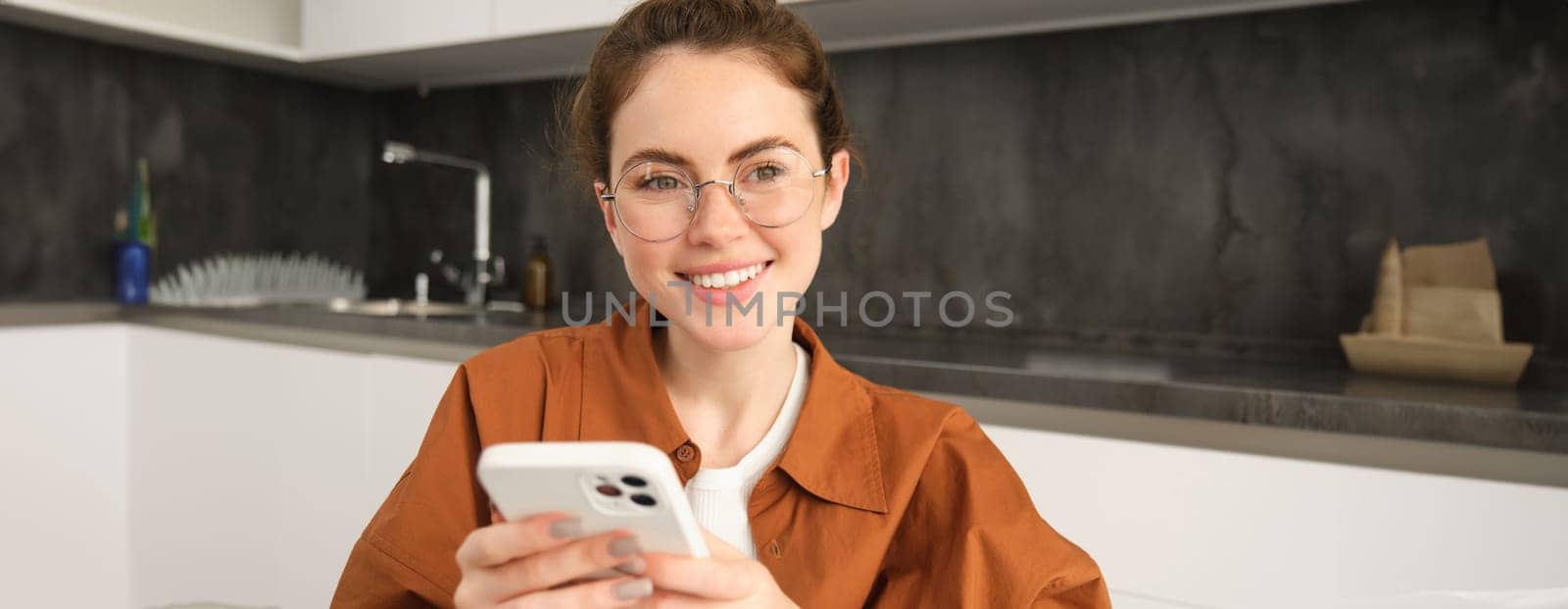 Close up portrait of smiling young woman, female model sitting in kitchen with smartphone, using mobile phone and smiling.