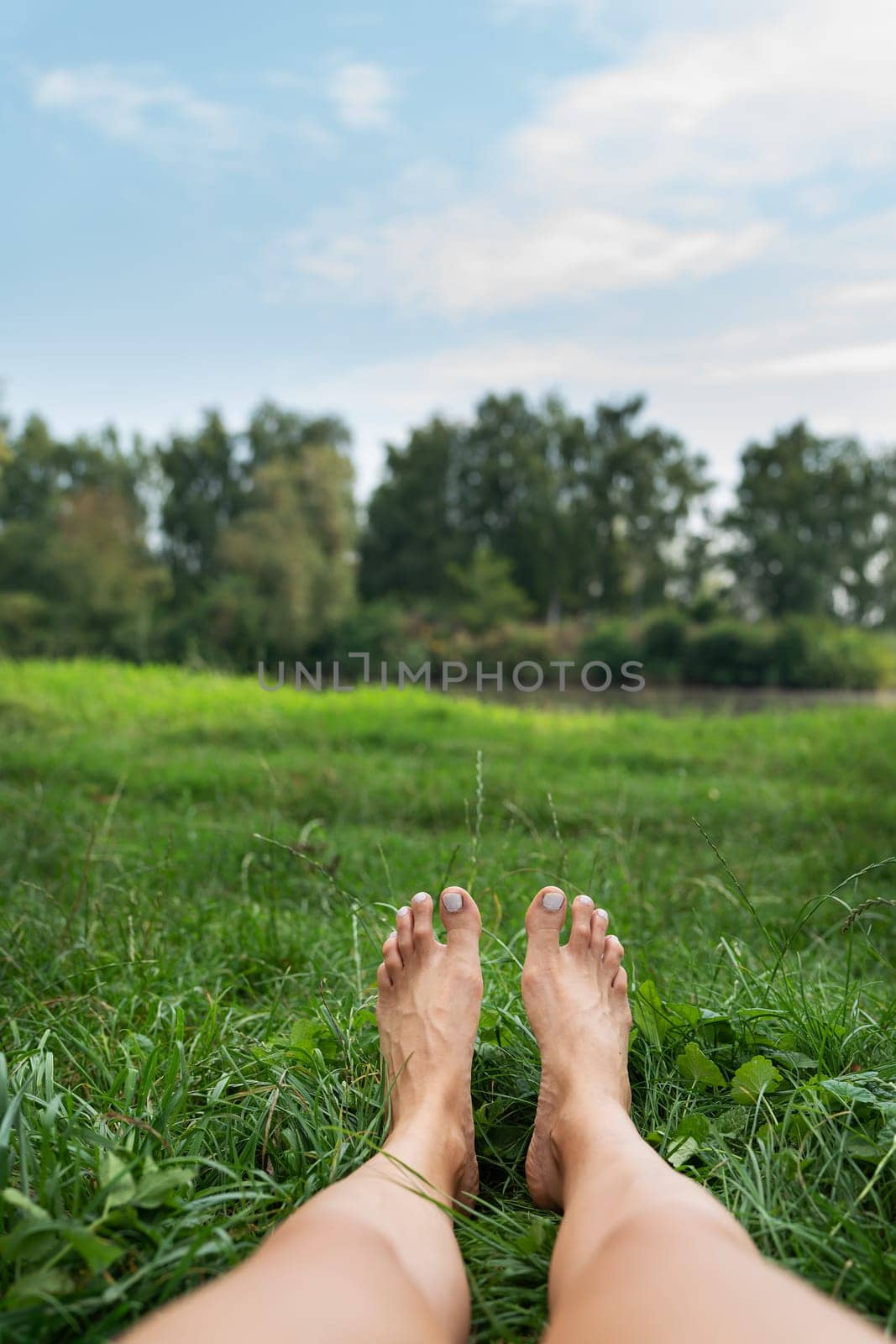 The image shows a pair of bare feet lying on a grassy field with a lake and a green park in the background. Toes point upward. The image is peaceful and calm