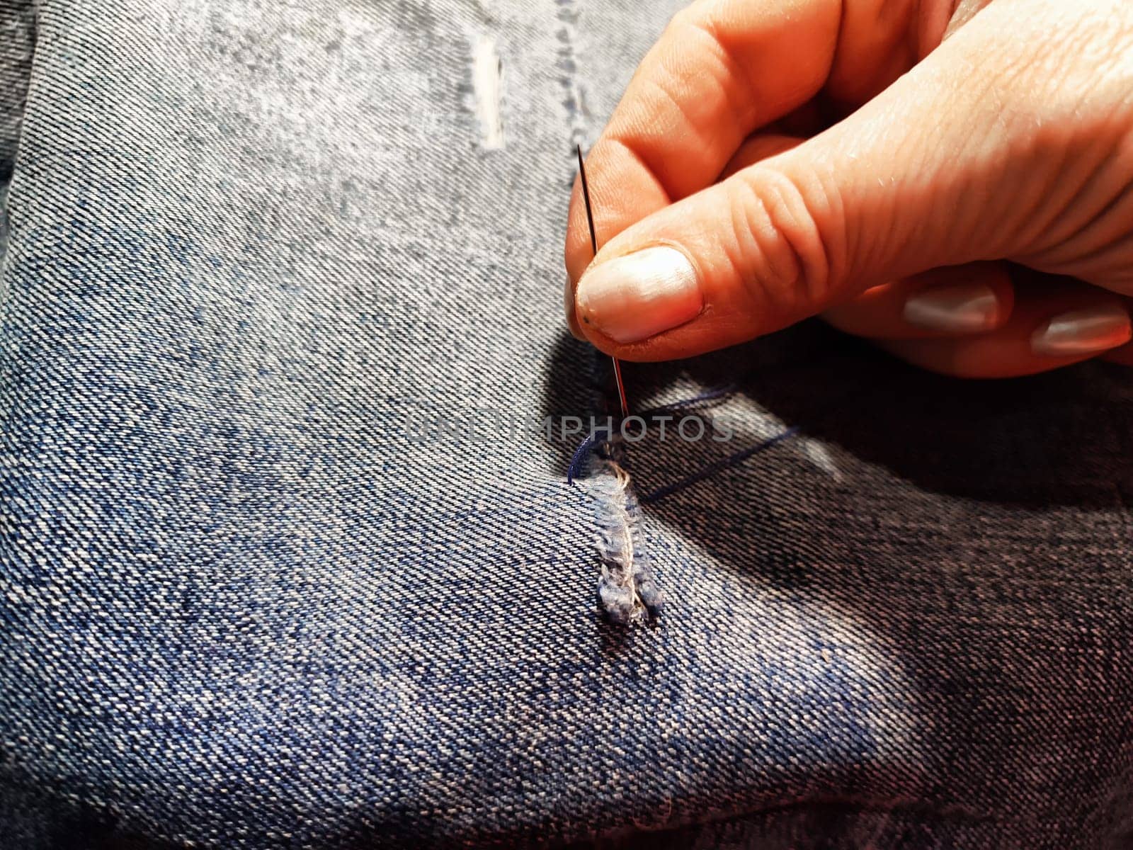 Woman repairing a pair of blue jeans. Fingers with a needle and torn jeans fabric. Sewing up a tear on blue fabric