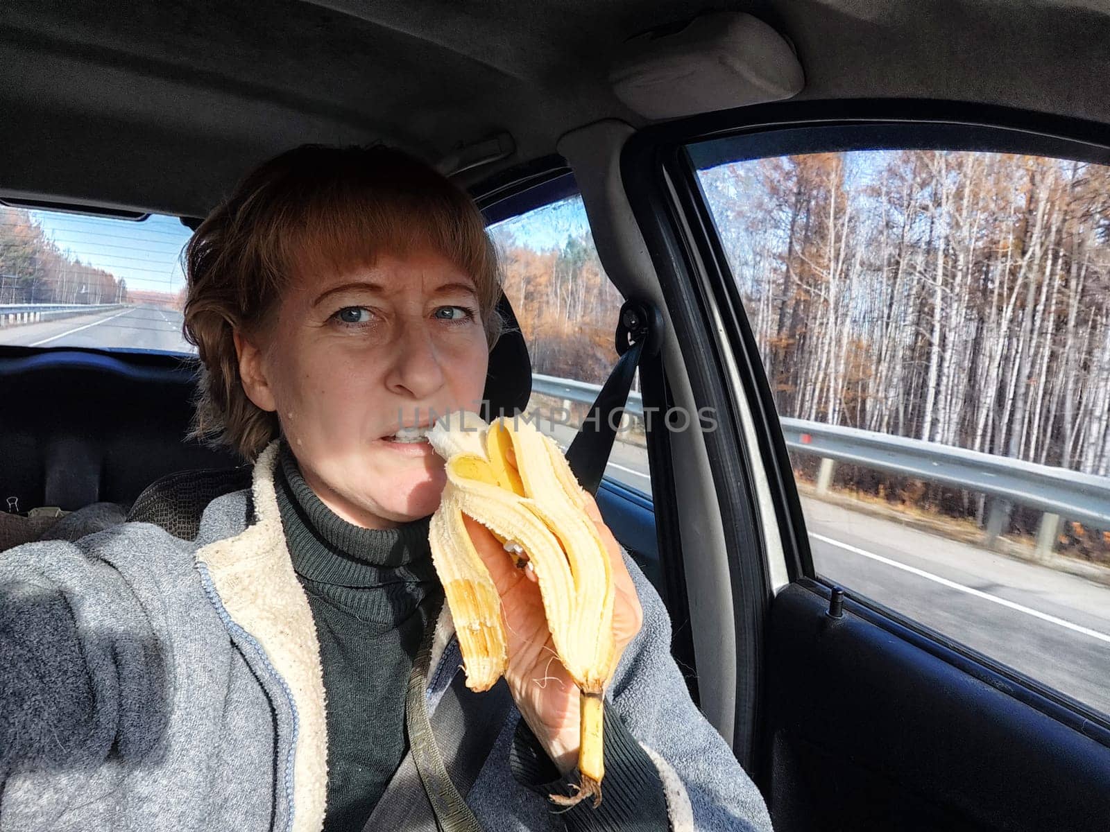 Portrait of female driver in solo journey. Adult mature middle aged woman holding steering wheel and banana. Eating while driving for to stay awake. Lady girl with food while travel or trip by car by keleny