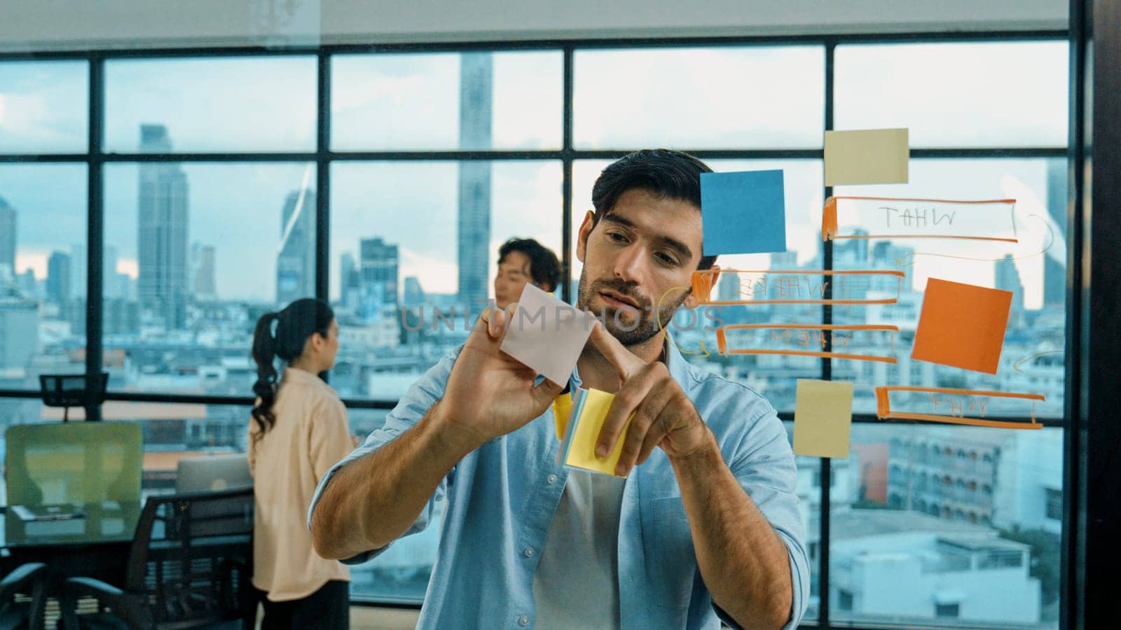 Smart project manager writing sticky notes and mind mapping at glass wall to planing strategy while multicultural businesspeople discussing about business plans at office with city view. Tracery