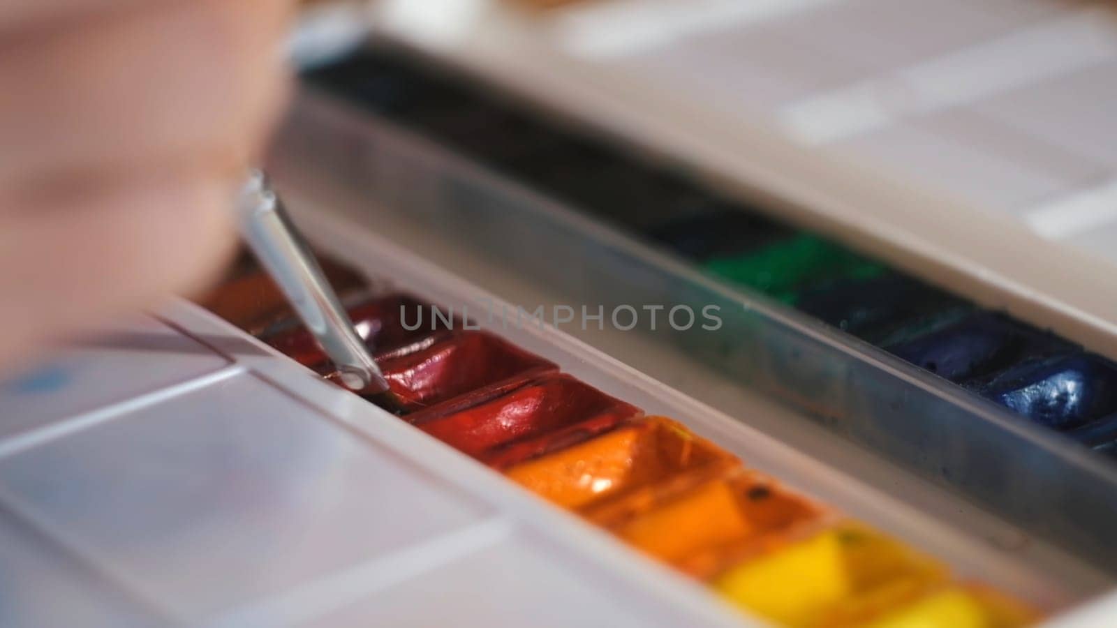 Putting paint brush into olorful oil paints. Close up of an artist hand holding a paint brush and mixing colors on clean white palette with water.