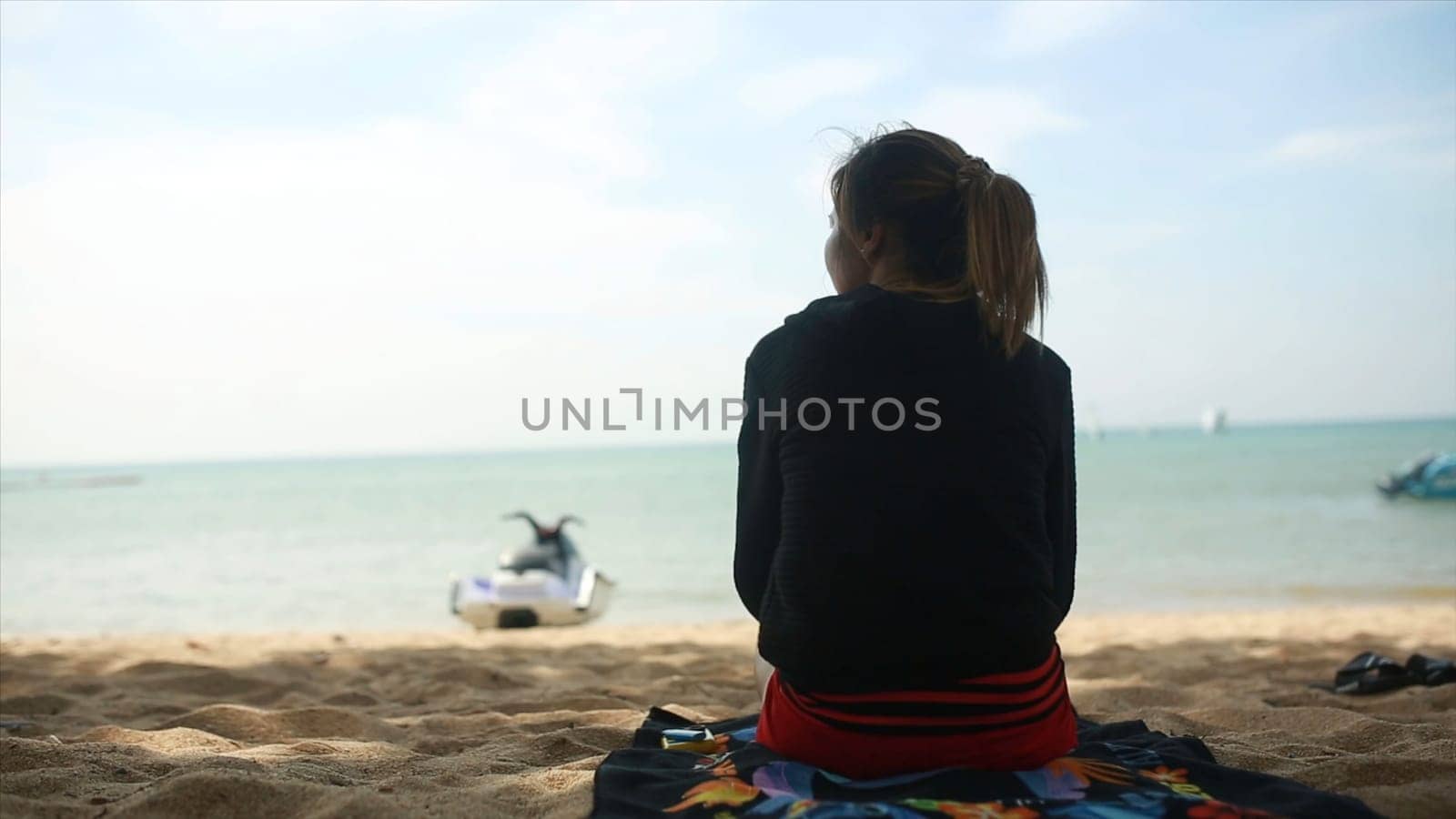 Rear view of a girl in a hoodie sitting on the beach and watch the sea, blue sky and sea background. Clip. Young woman in sportswear sitting on the beach.