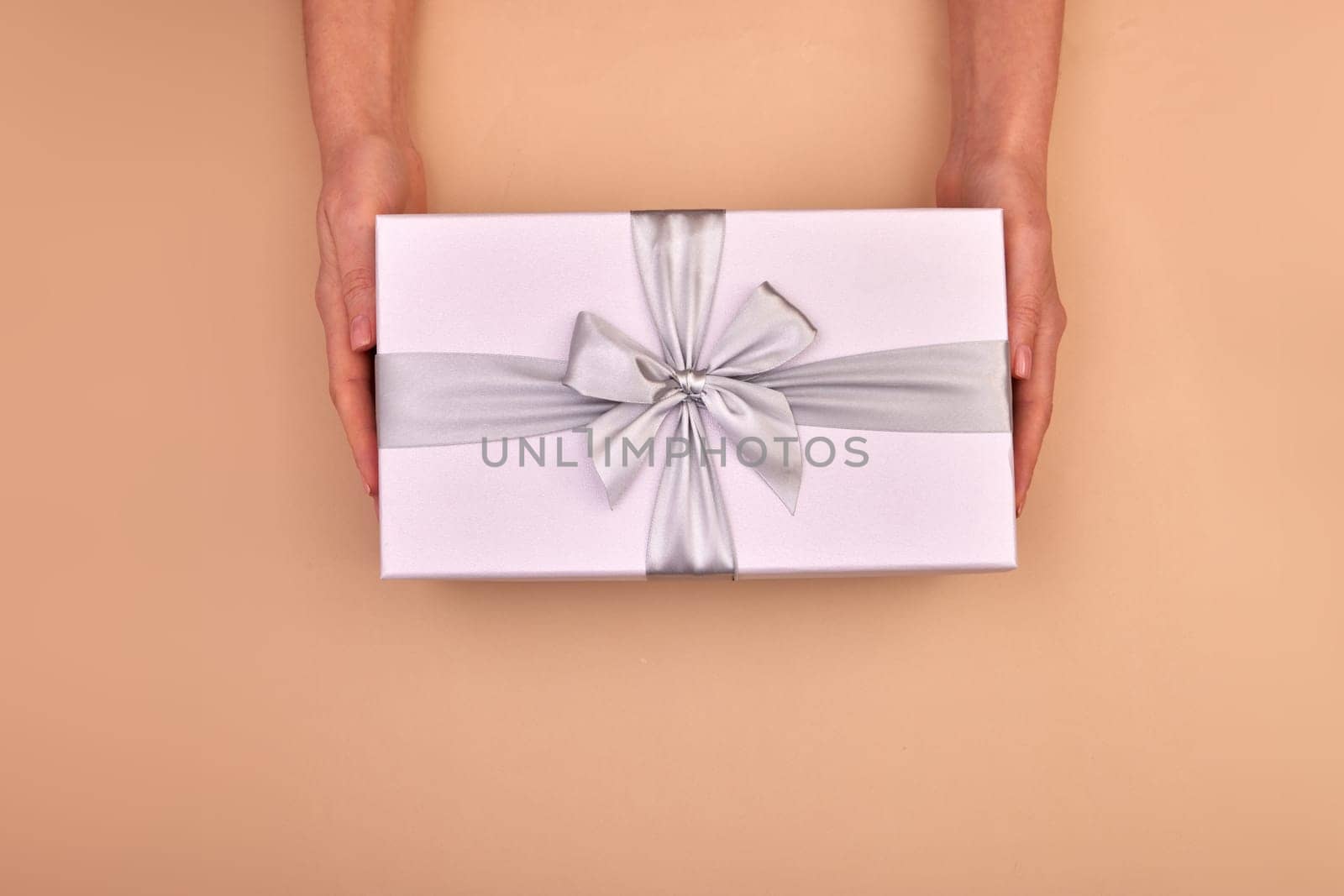 Female hands hold gray violet gift with ribbon bow on beige background flat lay top view copy space holiday present, Color of the Year for 2024