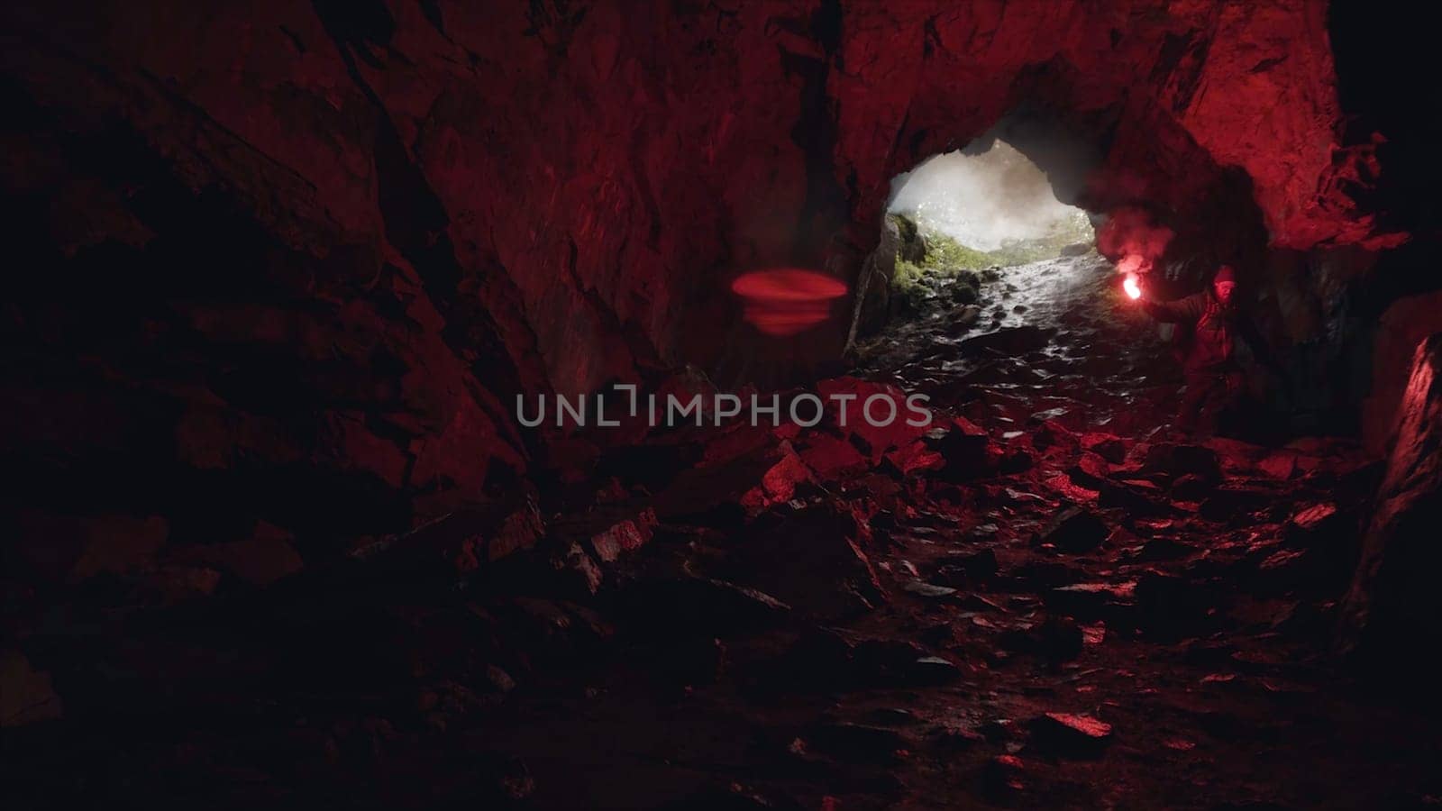 Man moving inside the dark and mysterious cavern with a red burning signal flare in his hand. Male explorer walking inside the cave.