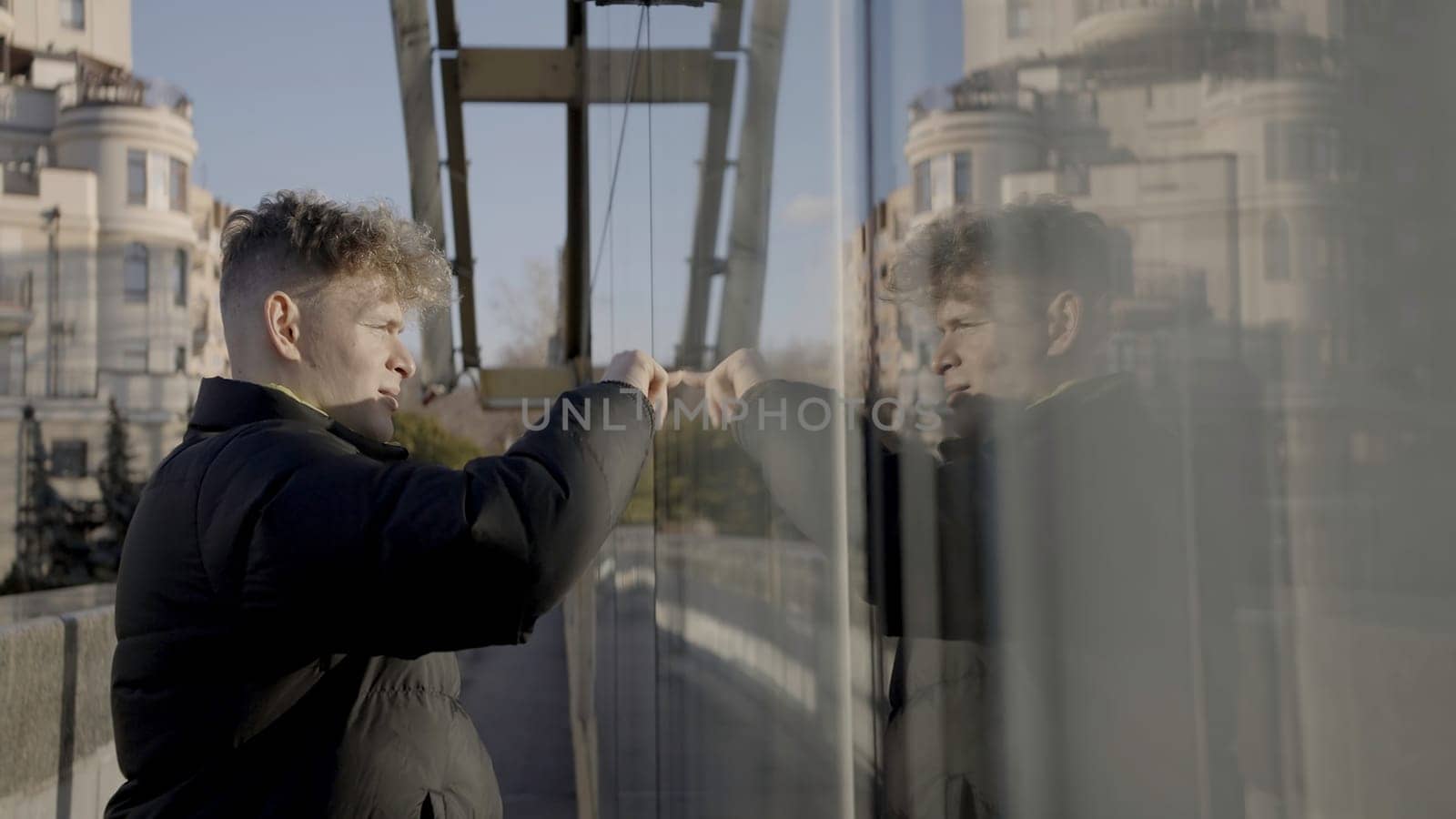 Young man draws on glass with finger. Action. Young attractive man draws on glass in city. Man runs finger along glass fence on sunny day.