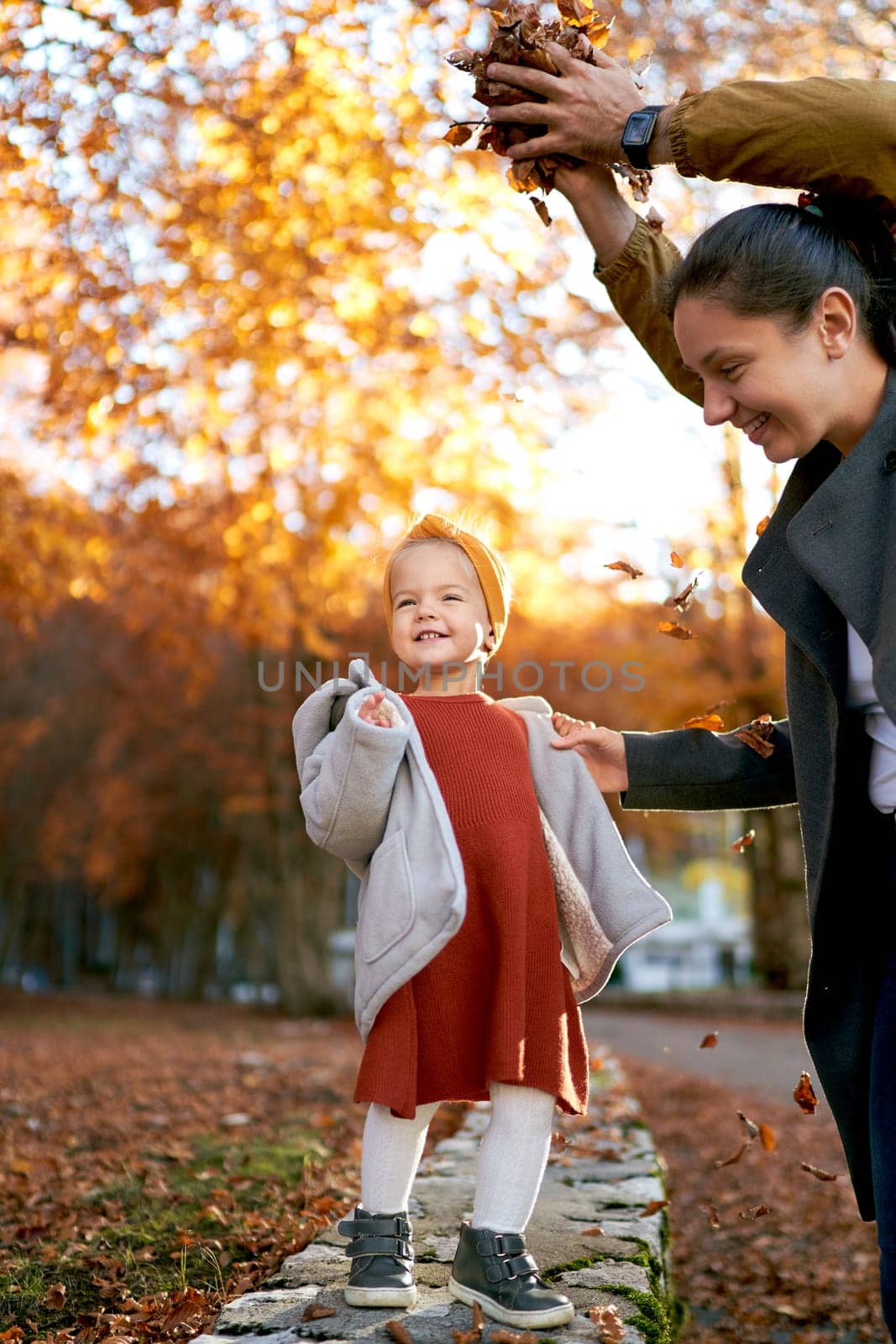 Dad stands with a handful of fallen leaves over mom standing with a little girl in the park by Nadtochiy