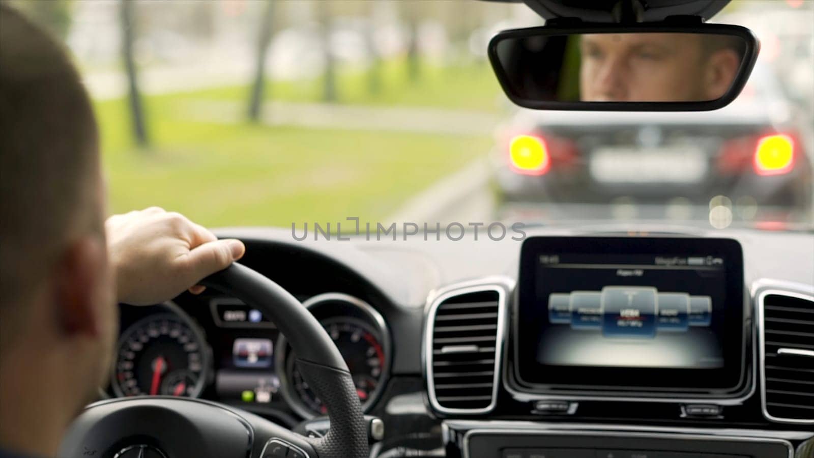 Man driving car from rear view in the city street along green trees alley. Man in grey shirt with black watch driving car on the road, modern car interior.