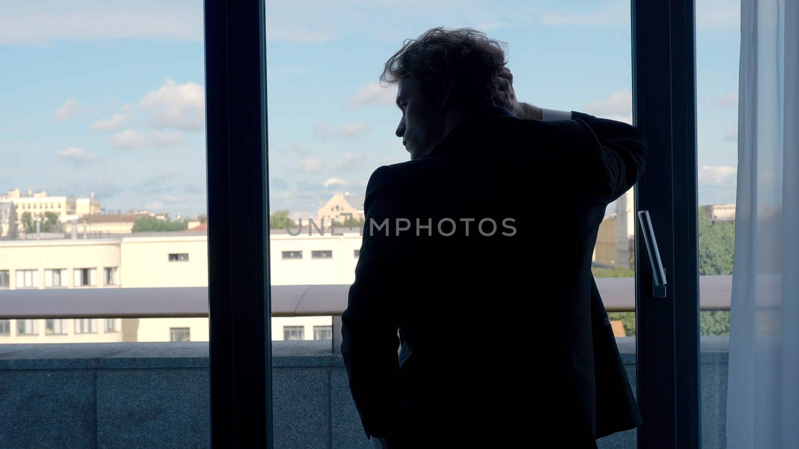 A man in a suit with long hair.Action. A handsome man stands at the window and looks at the city holding his hand behind his head, the sky is blue and small city houses are on the street