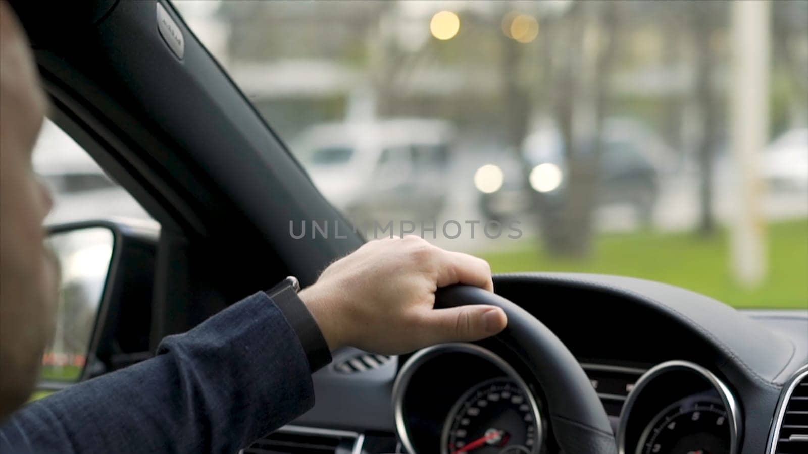 Man driving car from rear view in the city street along green trees alley. Man in grey shirt with black watch driving car on the road, modern car interior.