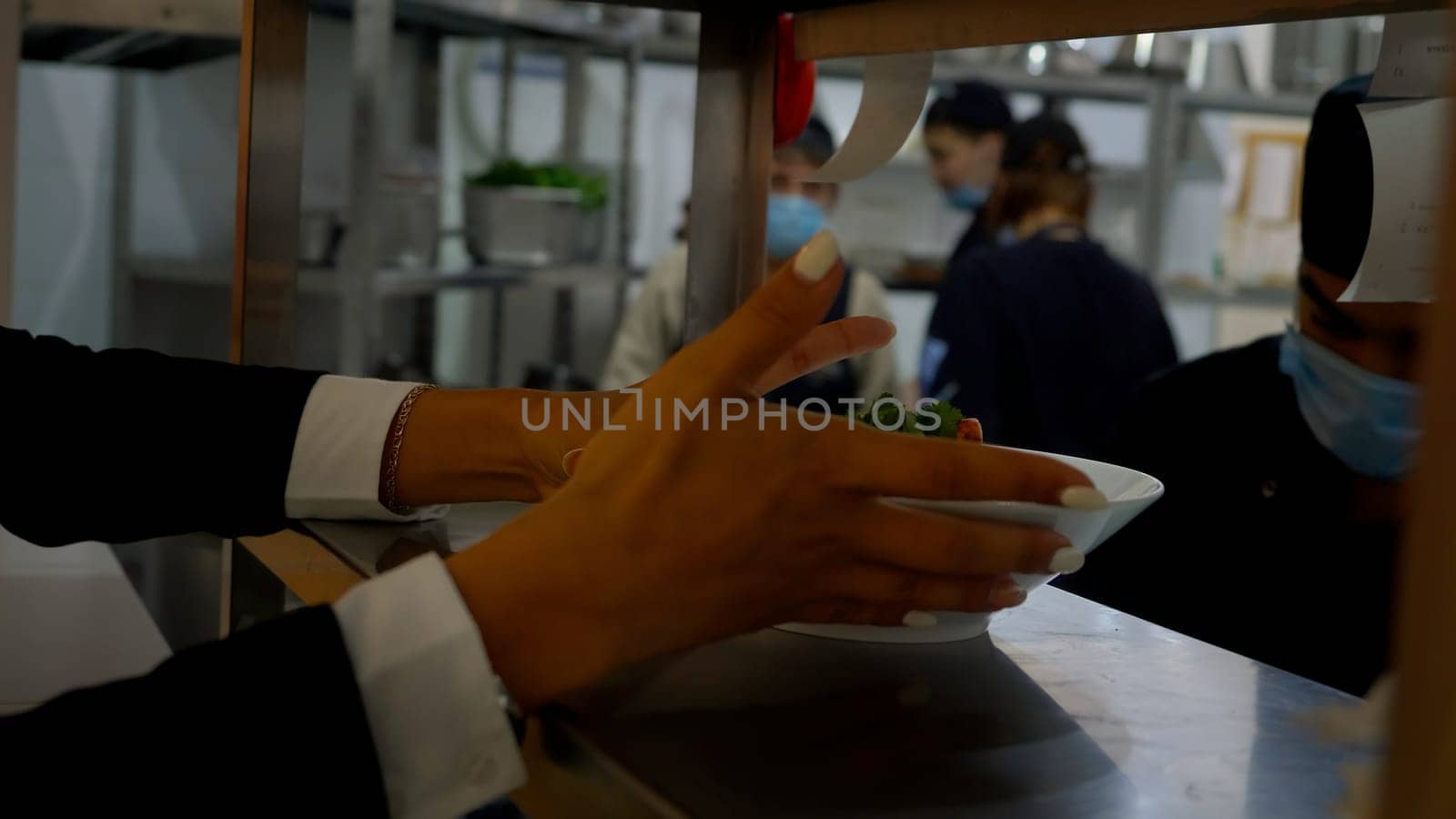 Germany - Berlin, December 15, 2023: In the restaurant, the waiter takes the prepared dish from the chef. Media. Plate with food on the metal surface at the cafe, cooks at work on the background
