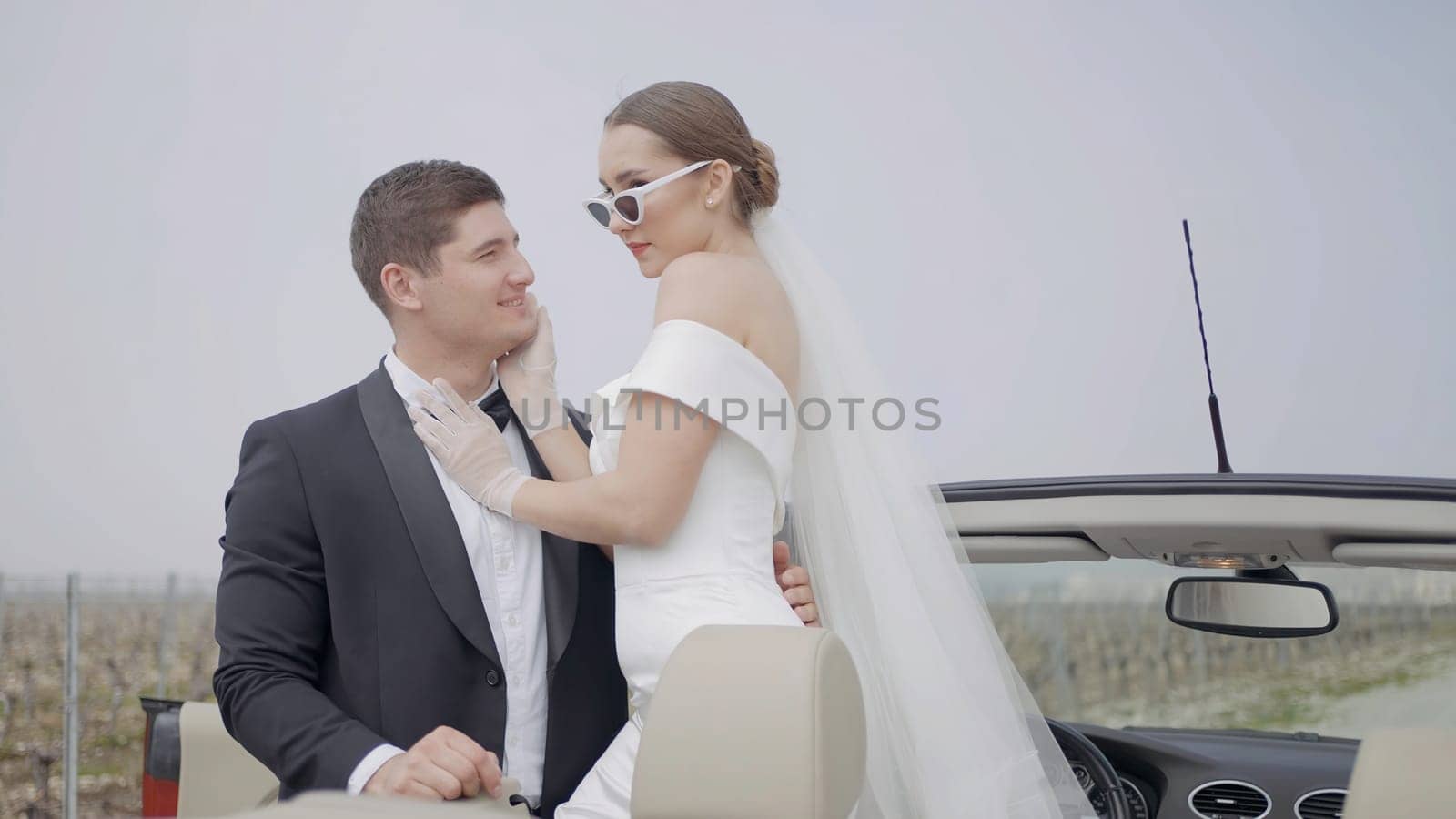 Just married couple, bride and groom in the open air. Action. Beautiful woman in white dress and sunglasses embracing man in suit while photosession in cabriolet