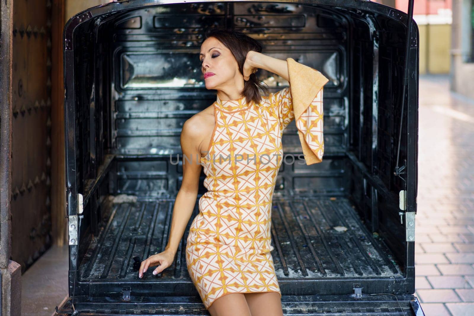 Sensual young woman in short gown with makeup, and eyeshadow standing stylishly with closed eye and hand on head in opened doorway at back of caravan on street